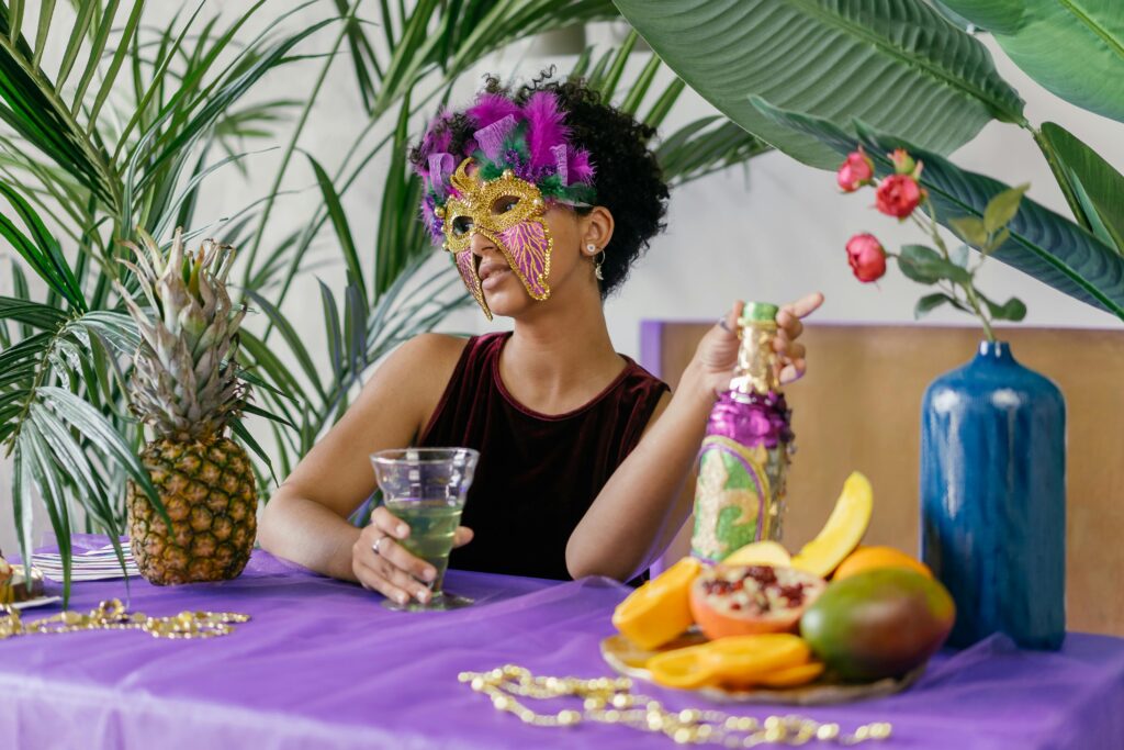 A woman in a vibrant masquerade mask enjoys tropical drinks surrounded by fresh fruits at an indoor party.
